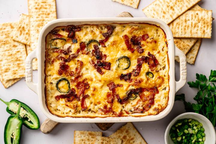 Creamy cheese dip in a baking dish, topped with jalapeños and crispy bacon, surrounded by crackers. Bright, appetizing, with fresh parsley nearby.